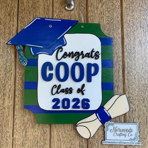 May include: A wooden graduation sign with a blue mortarboard, diploma, and the words "Congrats COOP Class of 2026." The sign is painted in shades of blue, green, and white, with a small business logo at the bottom.