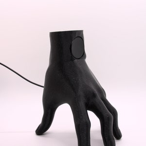 Puede incluir: Una l&aacute;mpara de mano negra impresa en 3D con un interruptor de luz redondo negro en la mu&ntilde;eca. La l&aacute;mpara est&aacute; sobre una superficie blanca.