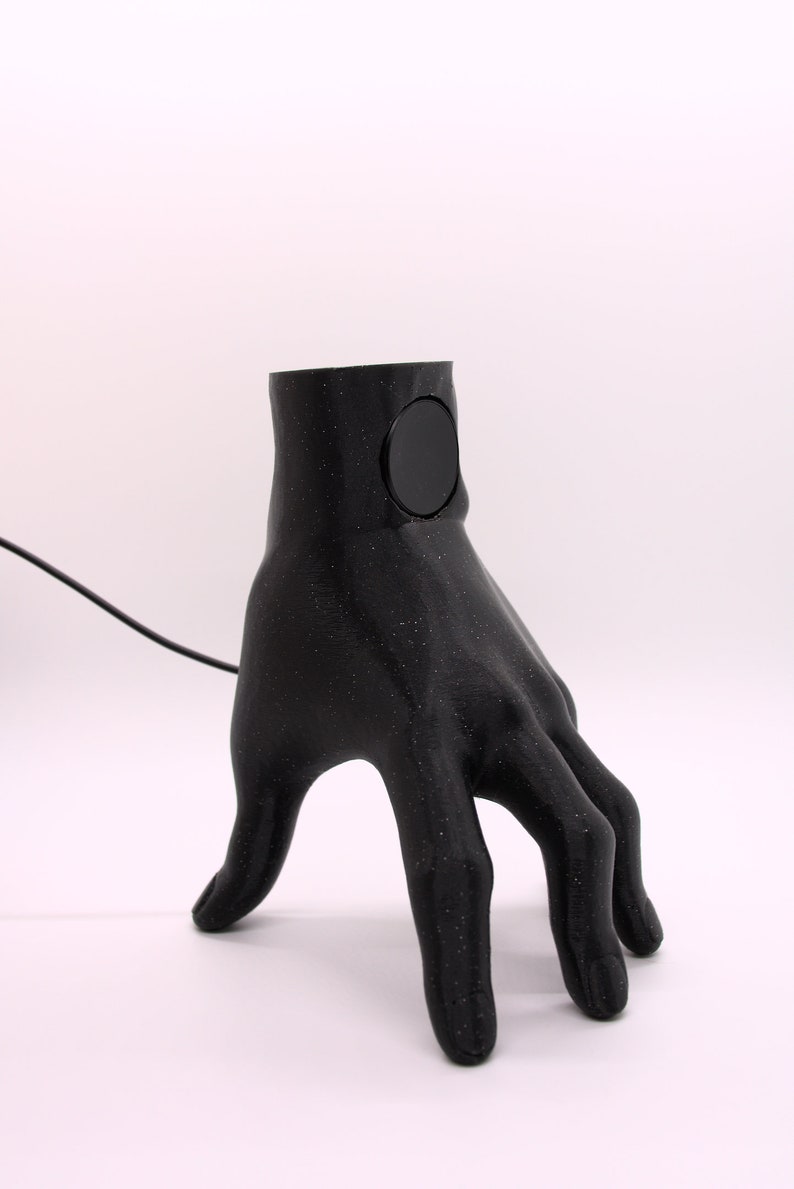 Puede incluir: Una l&aacute;mpara de mano negra impresa en 3D con un interruptor de luz redondo negro en la mu&ntilde;eca. La l&aacute;mpara est&aacute; sobre una superficie blanca.