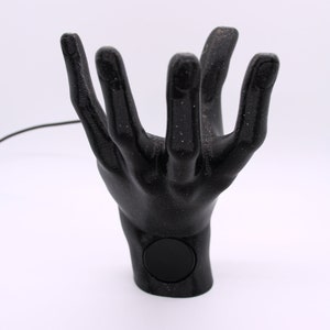Puede incluir: Un soporte para tel&eacute;fono de mano negro con una superficie texturizada y un recorte circular en la mu&ntilde;eca. El soporte est&aacute; hecho de pl&aacute;stico impreso en 3D.