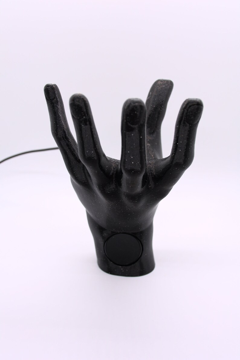 Puede incluir: Un soporte para tel&eacute;fono de mano negro con una superficie texturizada y un recorte circular en la mu&ntilde;eca. El soporte est&aacute; hecho de pl&aacute;stico impreso en 3D.