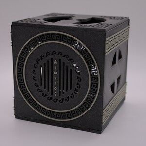 May include: A black cube-shaped box with a decorative silver circle on one side. The circle has a Greek key design around the edge and the word "AIGWIOS" written vertically in the center. The box has a small opening on each side.