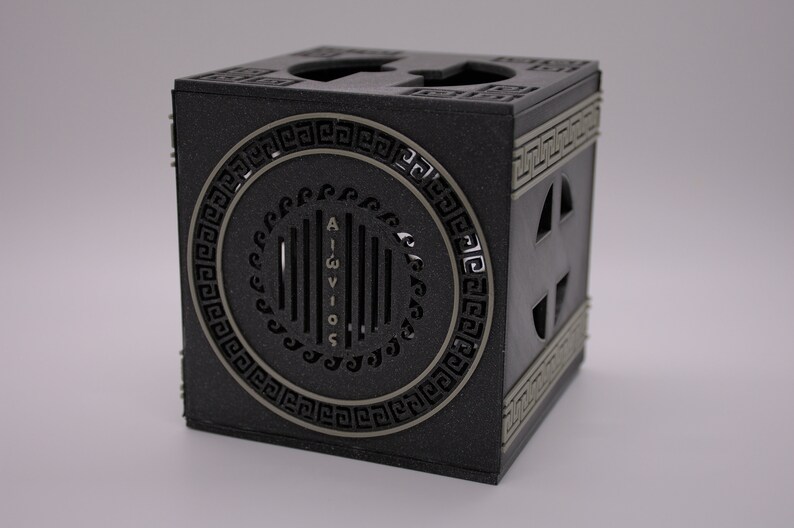 May include: A black cube-shaped box with a decorative silver circle on one side. The circle has a Greek key design around the edge and the word "AIGWIOS" written vertically in the center. The box has a small opening on each side.