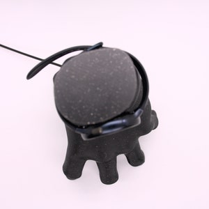 Puede incluir: Auriculares negros con una almohadilla texturizada y un cable negro. Los auriculares est&aacute;n colocados sobre un soporte negro con forma de mano.