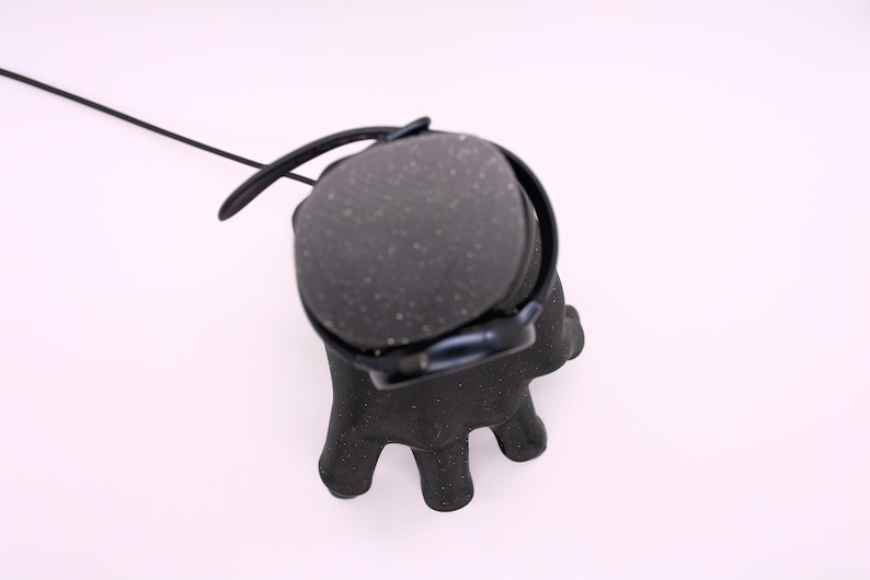 Puede incluir: Auriculares negros con una almohadilla texturizada y un cable negro. Los auriculares est&aacute;n colocados sobre un soporte negro con forma de mano.