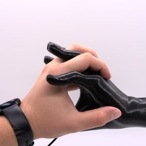 Puede incluir: Una mano negra impresa en 3D sosteniendo otra mano. La mano que sostiene la otra mano lleva un reloj de pulsera negro.