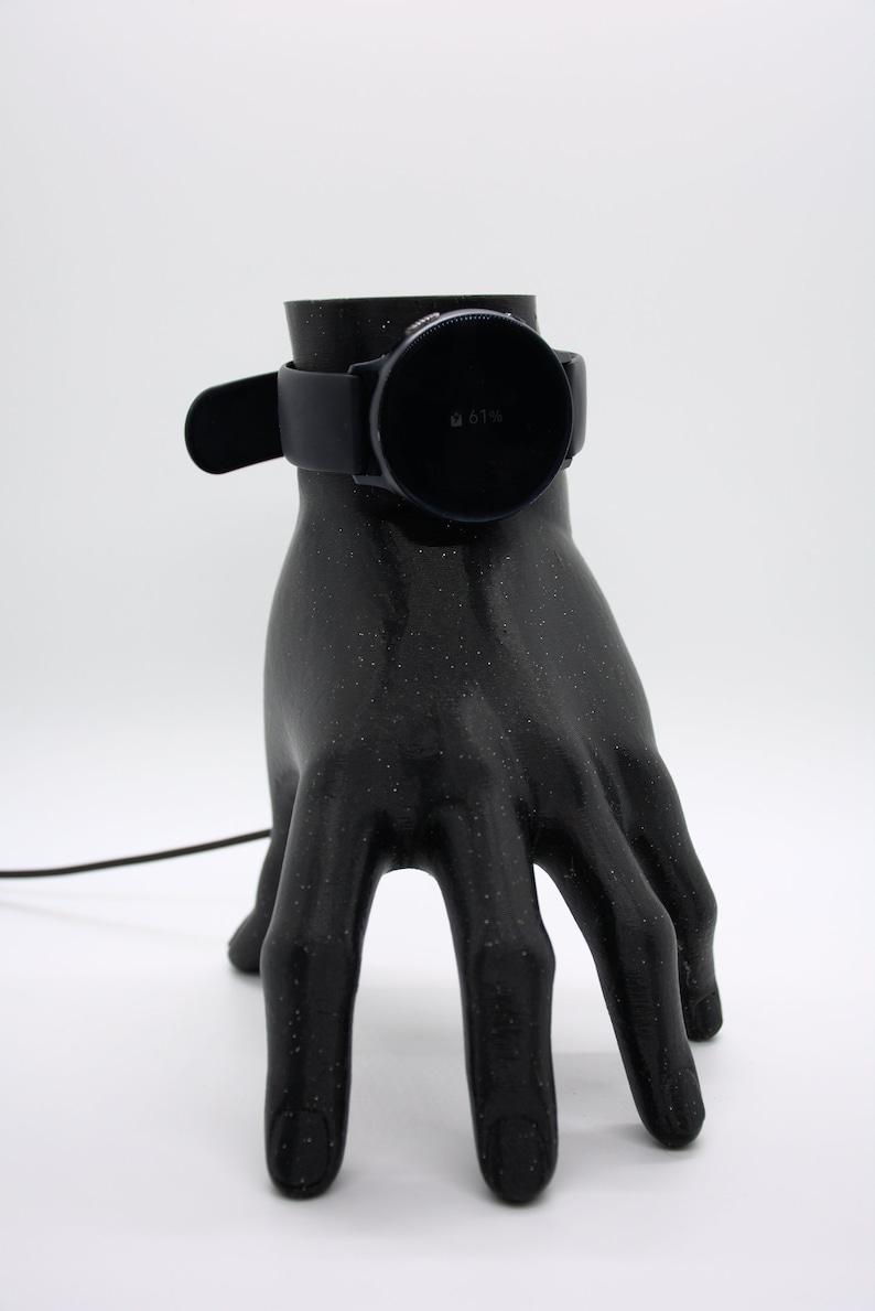 Puede incluir: Un soporte de carga impreso en 3D con forma de mano negra con un reloj inteligente negro en la parte superior. La pantalla del reloj inteligente muestra un 61% de bater&iacute;a.