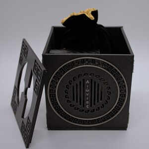 May include: A black wooden box with a gold design and a black velvet bag inside. The box has a Greek key pattern around the edges and a circular design on the front with the text "AIGWIOS".