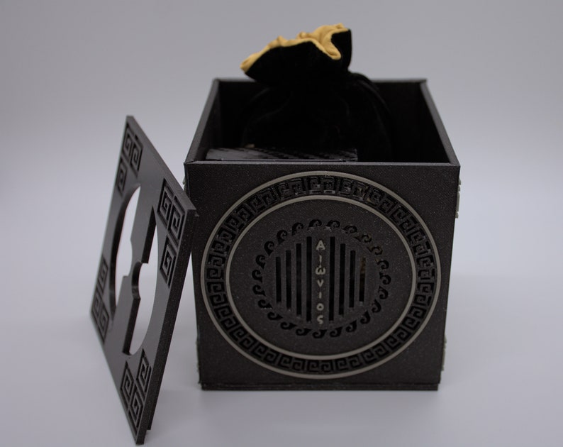 May include: A black wooden box with a gold design and a black velvet bag inside. The box has a Greek key pattern around the edges and a circular design on the front with the text "AIGWIOS".