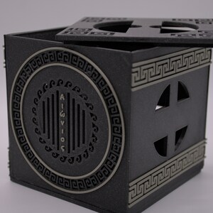 May include: A black cube-shaped box with a lid. The box has a decorative design with a circle containing the word "AIGVIVOS" and a Greek key pattern. The lid has a similar design with a cross-shaped cutout.