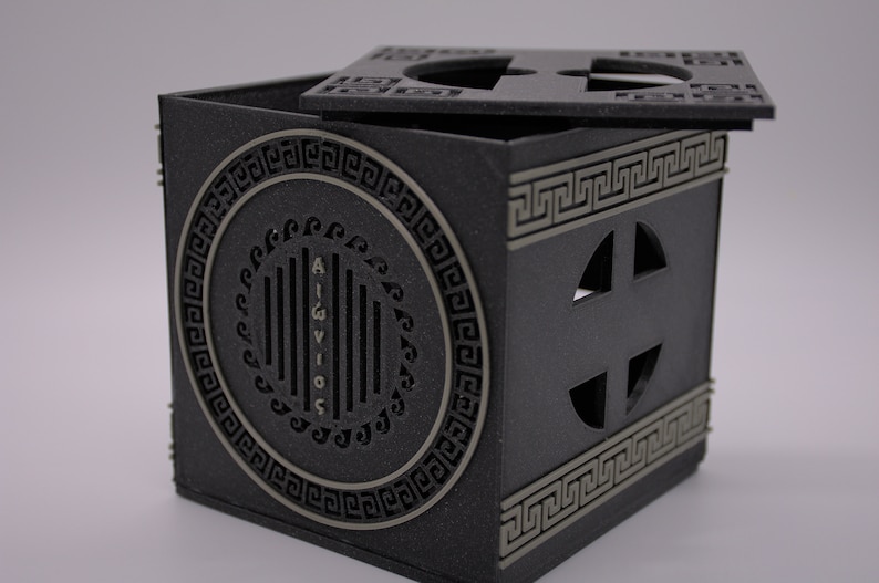 May include: A black cube-shaped box with a lid. The box has a decorative design with a circle containing the word "AIGVIVOS" and a Greek key pattern. The lid has a similar design with a cross-shaped cutout.