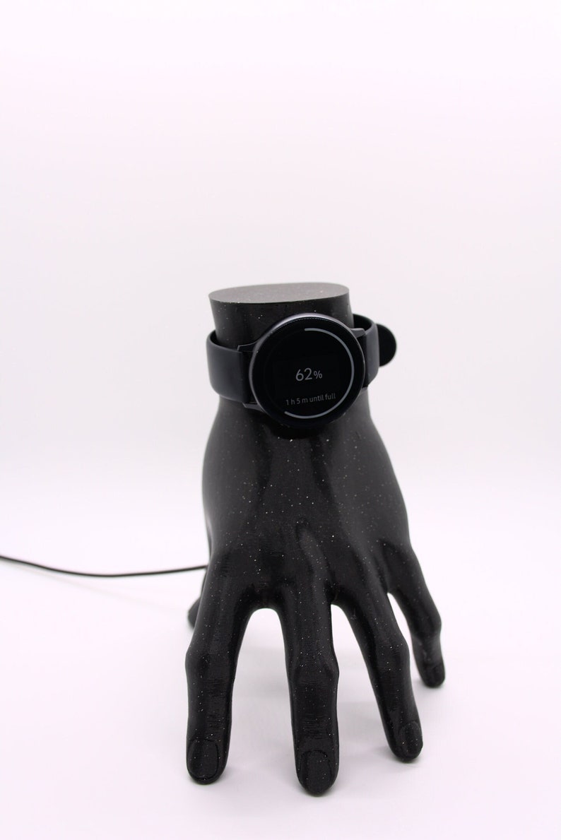 Puede incluir: Un reloj inteligente negro cargando en un soporte de mano impreso en 3D negro. La pantalla del reloj muestra 62% de bater&iacute;a y 1 h 57 m hasta que est&eacute; completamente cargado.