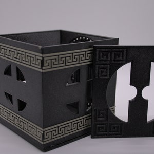 May include: A black square box with a Greek key design and a circular cutout. The box is open and the lid is lying next to it. The lid also has a circular cutout.