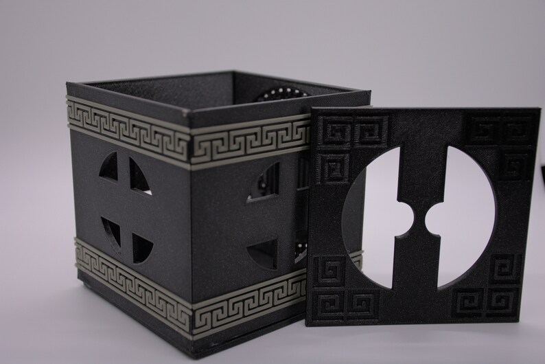 May include: A black square box with a Greek key design and a circular cutout. The box is open and the lid is lying next to it. The lid also has a circular cutout.