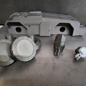 May include: A 3D printed model of a futuristic vehicle, disassembled into its component parts. The model is made of black plastic and includes a main body, four wheels, two guns, and a small engine.