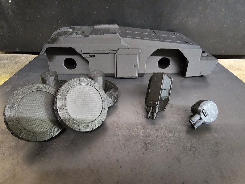 May include: A 3D printed model of a futuristic vehicle, disassembled into its component parts. The model is made of black plastic and includes a main body, four wheels, two guns, and a small engine.