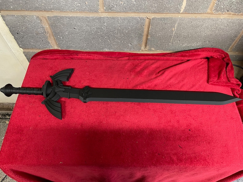 May include: A black plastic replica of the Master Sword from the Legend of Zelda video game series. The sword is lying on a red fabric surface.