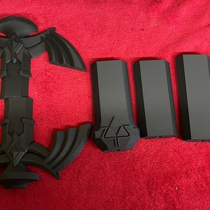 May include: A black plastic model of a sword, disassembled into multiple pieces. The sword has a triangular blade with a decorative design and a handle with a textured grip.