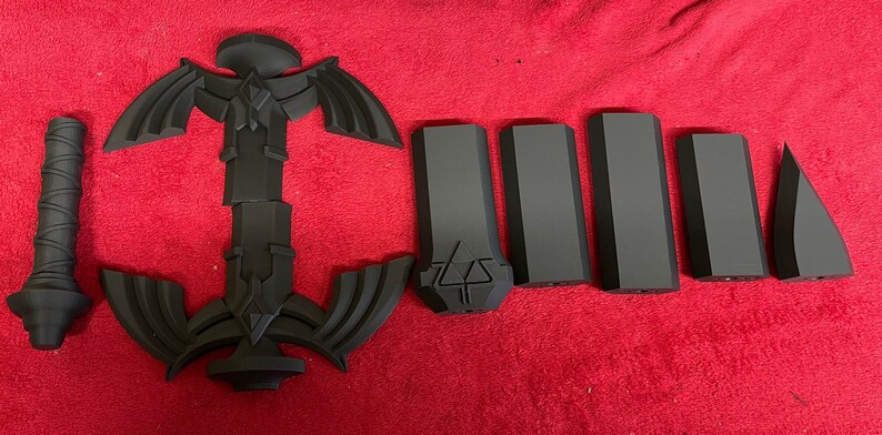 May include: A black plastic model of a sword, disassembled into multiple pieces. The sword has a triangular blade with a decorative design and a handle with a textured grip.