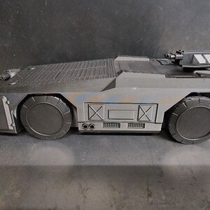 May include: A black toy military vehicle with a raised gun turret and two large wheels. The vehicle has a textured surface and a faded blue and brown paint job.