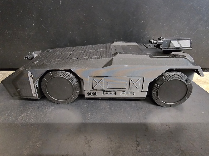 May include: A black toy military vehicle with a raised gun turret and two large wheels. The vehicle has a textured surface and a faded blue and brown paint job.