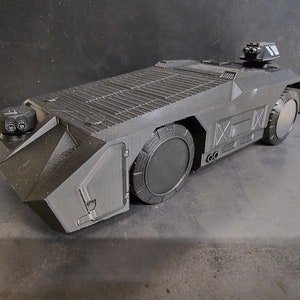 May include: A black, detailed, 3D printed model of a futuristic military vehicle with a raised platform and a turret on top.