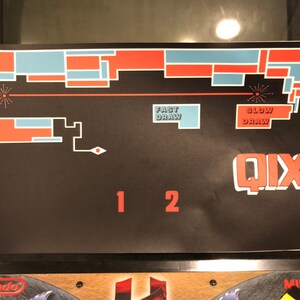 May include: A black and red game board with the word "QIX" in red letters. The board has a red and blue maze with the words "Fast Draw" and "Slow Draw" in white letters. The numbers "1" and "2" are in red letters.