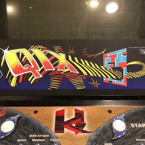 May include: A vintage arcade game cabinet with a black control panel and a colorful marquee featuring the word "QIX" in a 3D style. The marquee also includes a blue and yellow geometric pattern and the word "TAITO" in white.