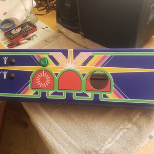 May include: A rectangular panel with a blue background, featuring a retro design with a sunburst and rainbow stripes. It has two black buttons, a green button, and three red and green arched sections. The panel is likely for an arcade machine.