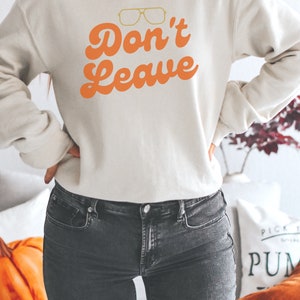 May include: Cream-colored sweatshirt with the text "Don't Leave" in orange, along with a graphic of glasses. The sweatshirt is paired with dark gray jeans. The overall theme is casual wear.
