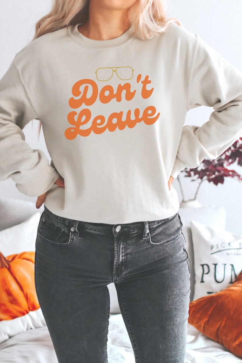 May include: Cream-colored sweatshirt with the text "Don't Leave" in orange, along with a graphic of glasses. The sweatshirt is paired with dark gray jeans. The overall theme is casual wear.