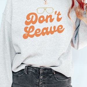 May include: A light gray sweatshirt with the words "Don't Leave" in orange script, along with a pair of gold-colored glasses. The sweatshirt is paired with dark gray jeans.