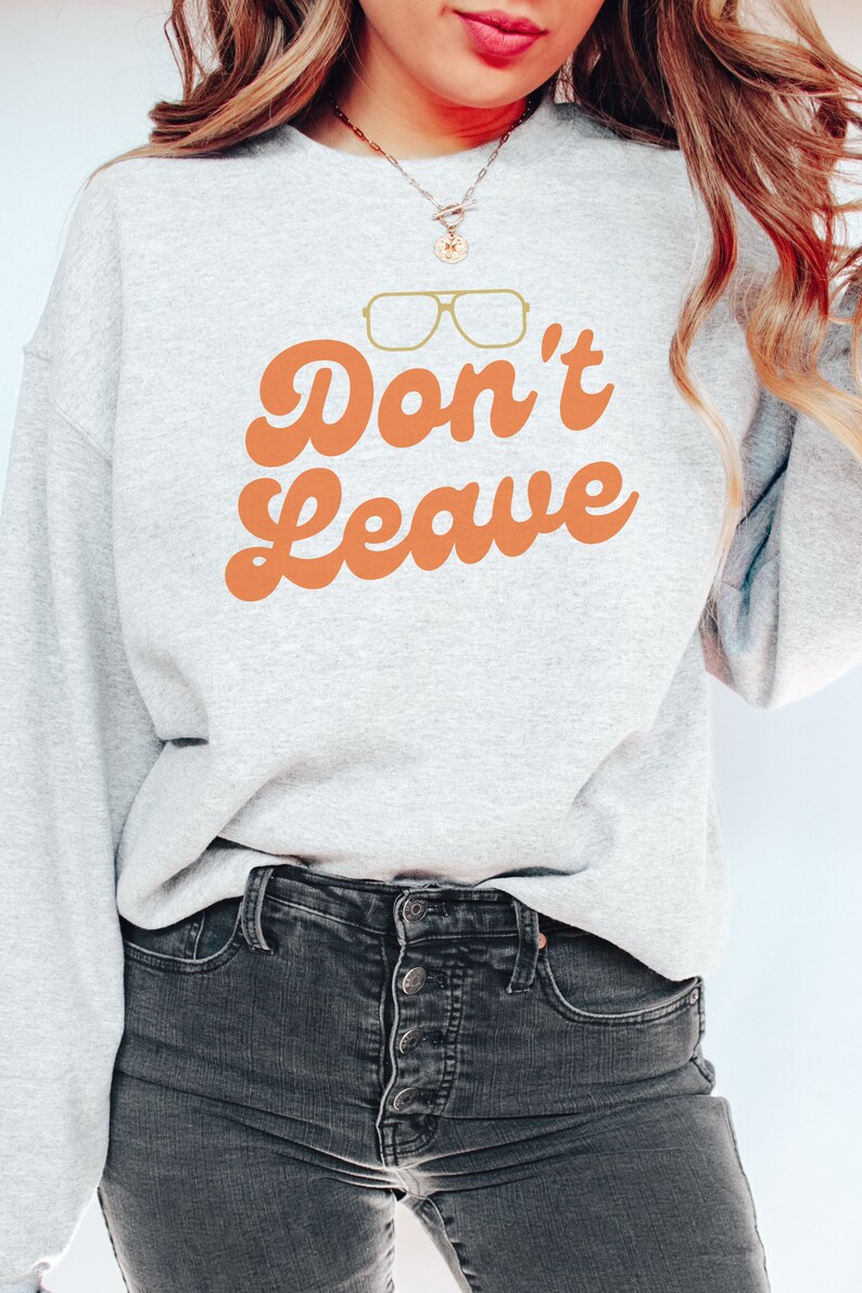 May include: A light gray sweatshirt with the words "Don't Leave" in orange script, along with a pair of gold-colored glasses. The sweatshirt is paired with dark gray jeans.