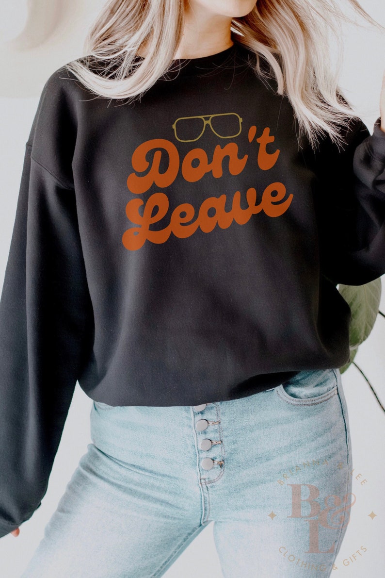May include: Black sweatshirt with the text "Don't Leave" in orange script and a pair of gold-rimmed glasses above the text. The sweatshirt is paired with light blue jeans.
