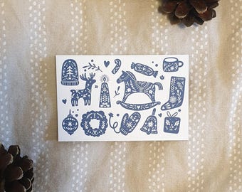 May include: A white card featuring blue Christmas-themed illustrations. The card includes images of a reindeer, rocking horse, ornaments, and other festive elements. Two pine cones are in the background.