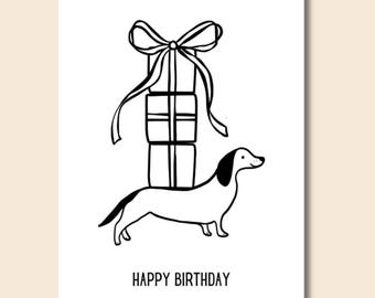 May include: Black and white illustration of a dachshund carrying a stack of wrapped presents with a bow on top. The text "Happy Birthday" is below the dog.
