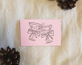 May include: A pink card with a black illustration of two champagne glasses clinking, each adorned with a bow. The card is set on a beige fabric with white polka dots, accompanied by two pine cones.