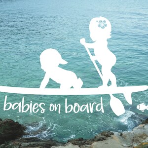 May include: White silhouette of two babies on a surfboard with the text "babies on board" below. The surfboard is on top of blue water with a rocky shore in the background.