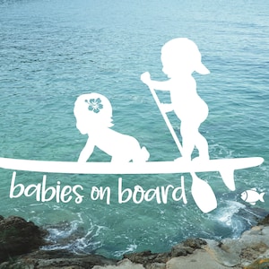 May include: White silhouette of two babies on a surfboard with the text "babies on board" in a cursive font. The background is a blue ocean with a rocky shore.