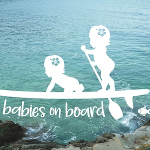 May include: White silhouette of two babies on a surfboard with the text "babies on board" in white. The background is a blue ocean with a rocky shore.