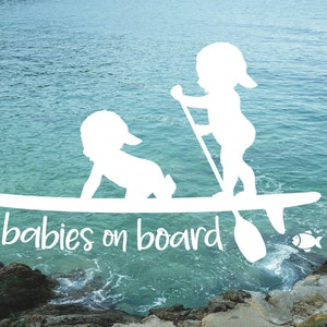 May include: White silhouette of two babies on a surfboard with the text "babies on board" in white. The background is a blue ocean with a rocky shore.