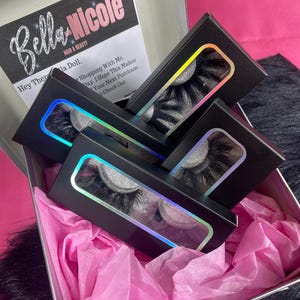 May include: A set of four black eyelash boxes with a holographic rainbow effect. The boxes are arranged in a stack, with the top box open to reveal a pair of black eyelashes. The boxes are sitting on pink tissue paper inside a silver box.