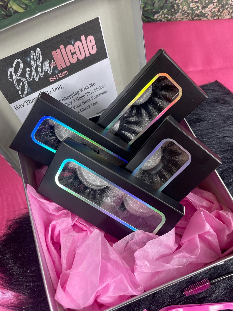 May include: A set of four black eyelash boxes with a holographic rainbow effect. The boxes are arranged in a stack, with the top box open to reveal a pair of black eyelashes. The boxes are sitting on pink tissue paper inside a silver box.