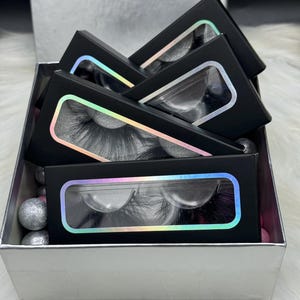 May include: A silver gift box with a black lid, containing multiple black boxes with clear windows displaying false eyelashes. The boxes are stacked on top of each other and have a holographic iridescent border around the window. The text "Bella Nicole" is printed on the lid of the box.