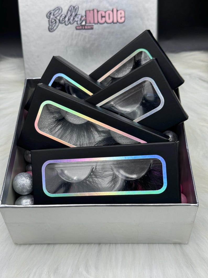May include: A silver gift box with a black lid, containing multiple black boxes with clear windows displaying false eyelashes. The boxes are stacked on top of each other and have a holographic iridescent border around the window. The text "Bella Nicole" is printed on the lid of the box.
