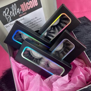 May include: A set of four black eyelash boxes with iridescent holographic accents. The boxes are stacked on top of each other and are sitting in a pink tissue-lined gift box.