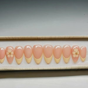 May include: A set of almond-shaped press-on nails. Each nail has a pale pink base with a light yellow French tip. Two nails feature a white flower design with gold accents. The nails are arranged in a rectangular box.