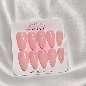 May include: A set of ten almond-shaped press-on nails in a solid light pink color with a glossy finish. The nails are displayed on a white rectangular card labeled Collection Nail Art with size checkboxes for XS, S, M, and L at the bottom.