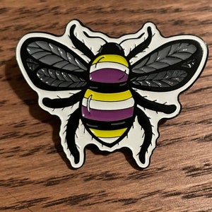 May include: A black and white enamel pin of a bee with yellow and purple stripes on its abdomen. The bee has black wings and is set against a white background.