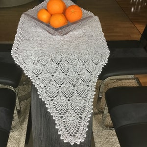 Puede incluir: Un camino de mesa de crochet blanco con un patrón delicado, colocado sobre una mesa de vidrio con un tazón de naranjas en el centro.